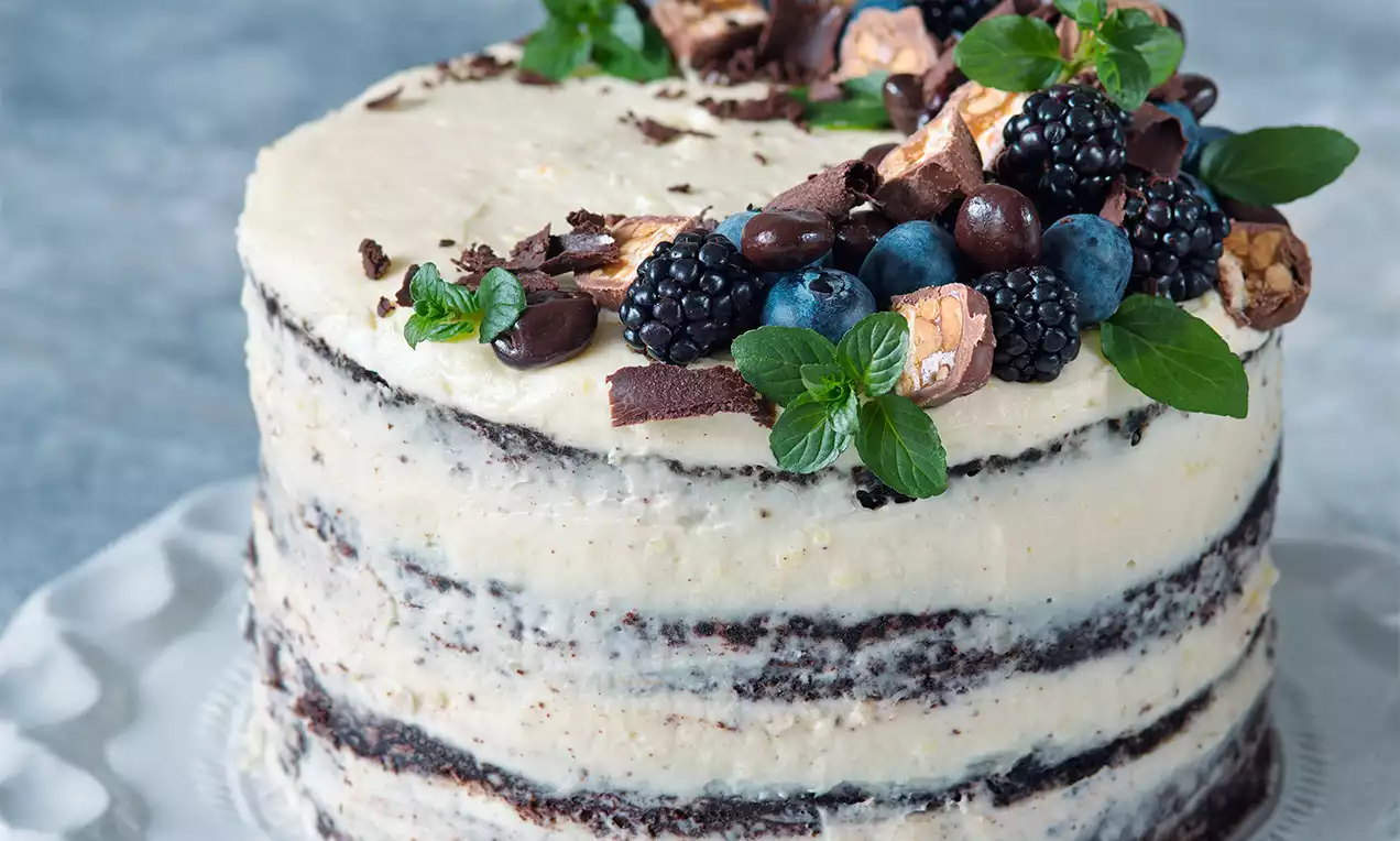 Čokoládový tort s vrstvami tmavej kakaovej piškóty a svetlej krémovej náplne, ozdobený čerstvým ovocím, čokoládou a mätovými lístkami na bielom tanieri