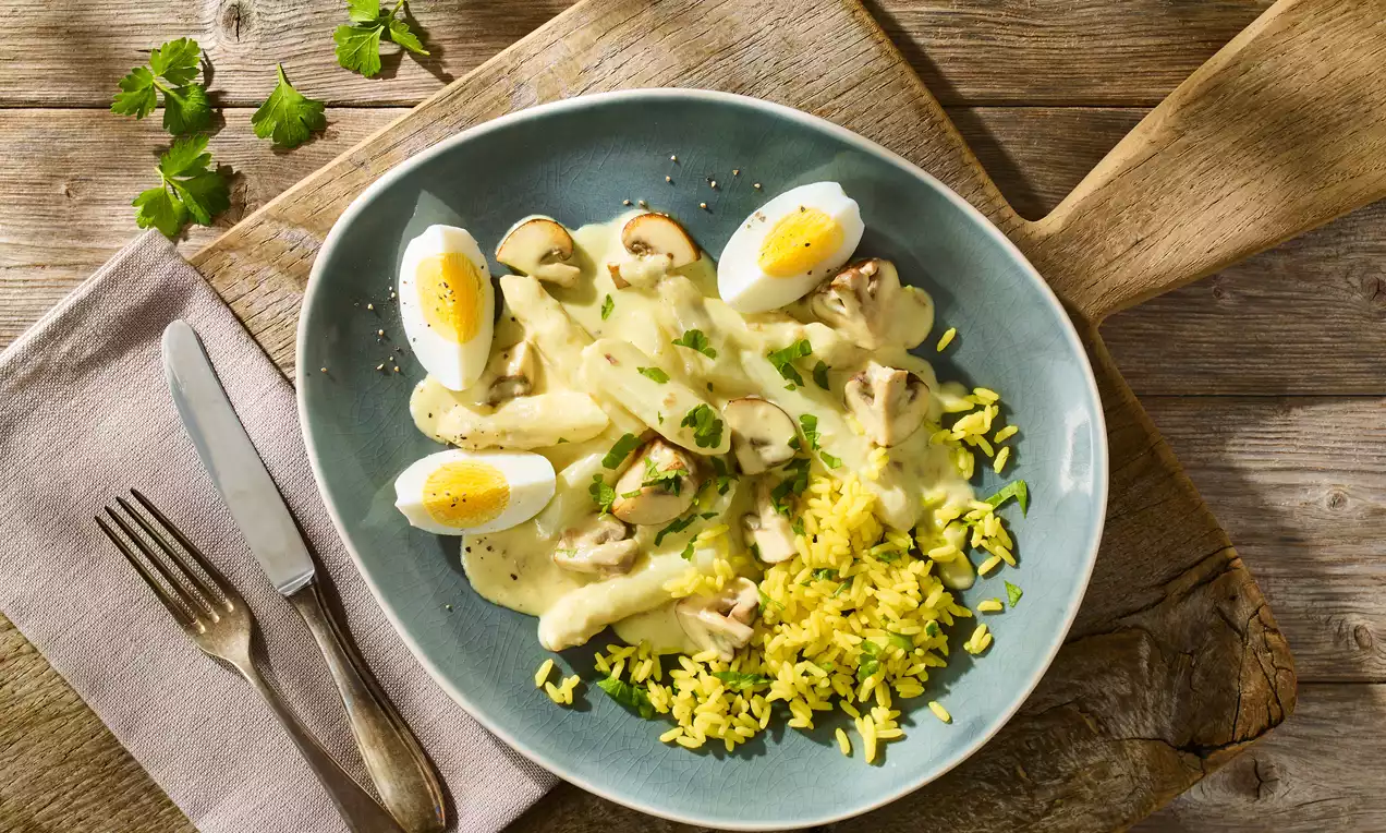 Spargelfrikassee mit weißen Spargelstangen, Champignons, Eierhälften und gelbem Reis auf hellblauem Teller, garniert mit Petersilie