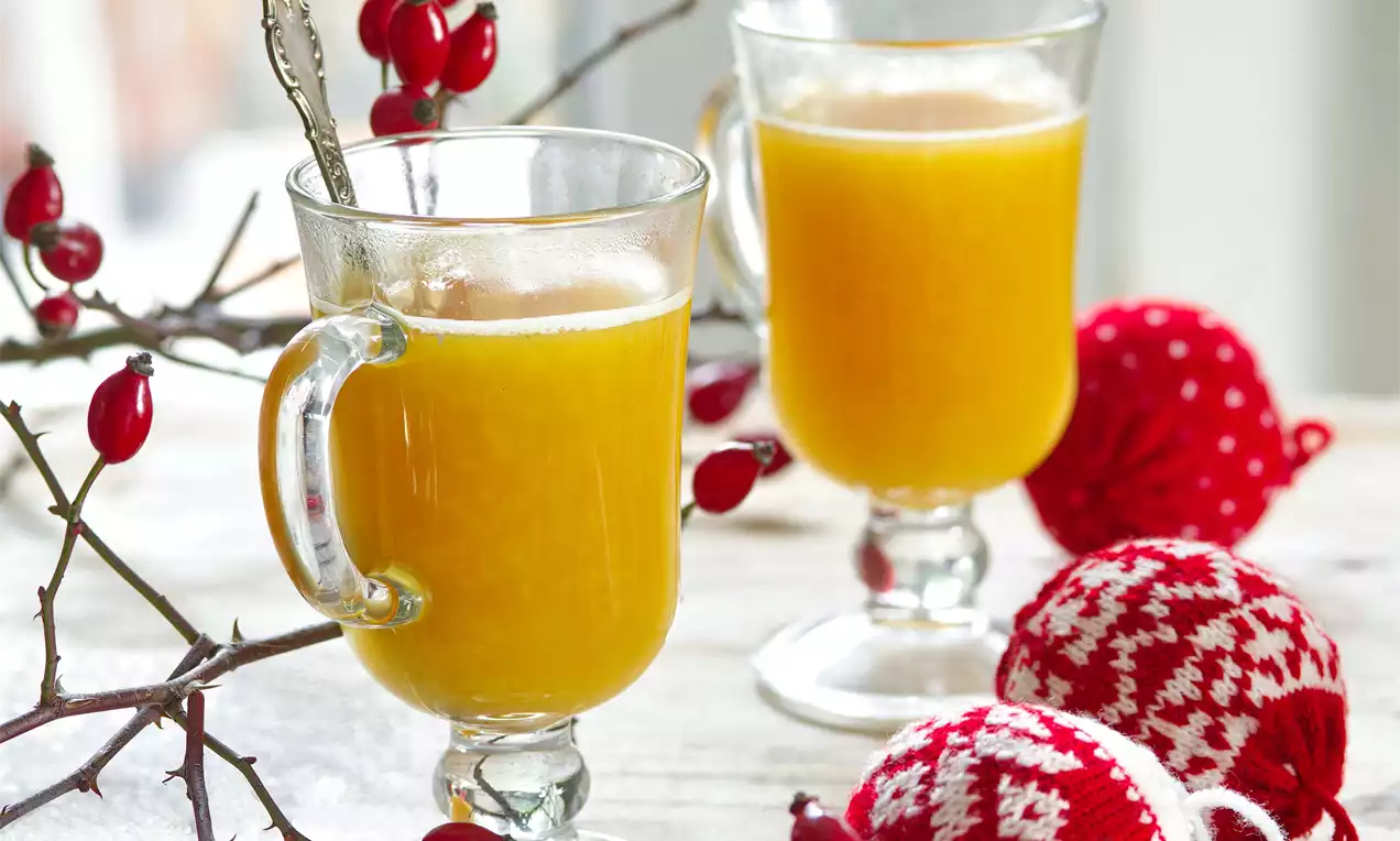 Hřejivý šípkový punč jasně oranžové barvy v průhledné sklenici s uchem, obklopený větvičkami šípků a pletenými vánočními ozdobami