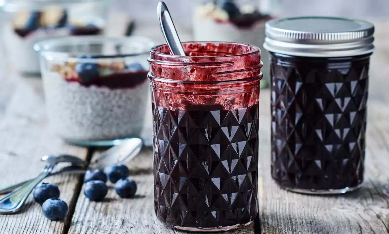 Bøtter med mørkelilla blåbærsyltetøj i facetteret glas, den ene med ske, omgivet af friske blåbær og chia-dessert i baggrunden