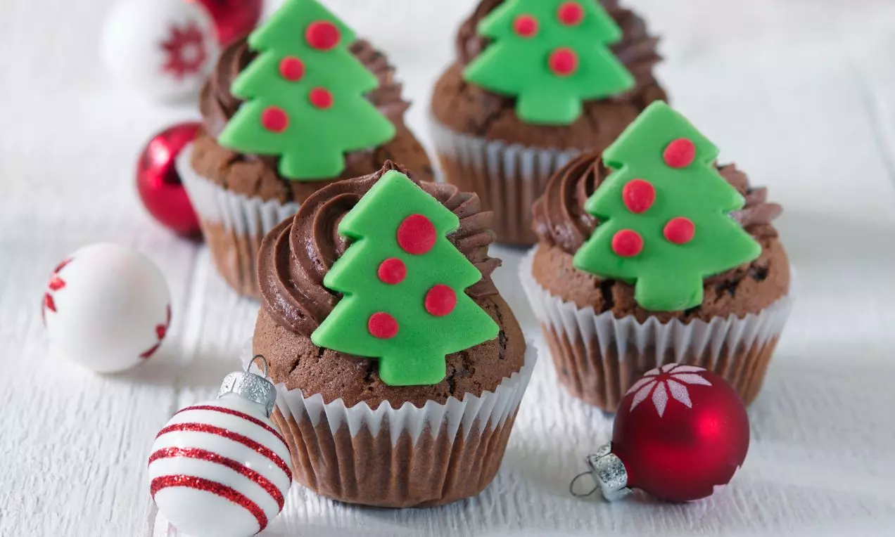 Čokoládové cupcakes s nadýchaným hnědým krémem, ozdobené zeleným cukrovým stromečkem s červenými tečkami, obklopené vánočními baňkami