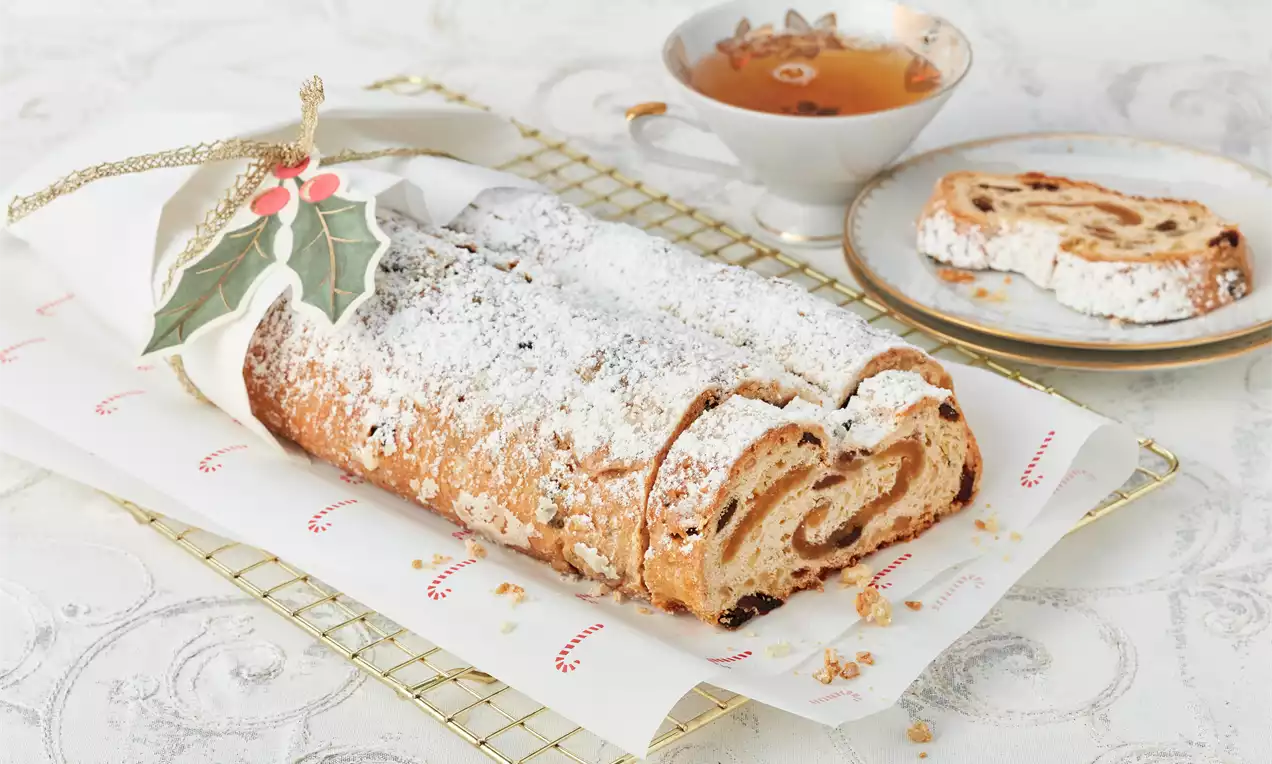 Goldbrauner Marzipanstollen mit hellen Zuckerschichten, locker geschnitten auf weißem Papier, dekoriert mit grünem Stechpalmenschmuck