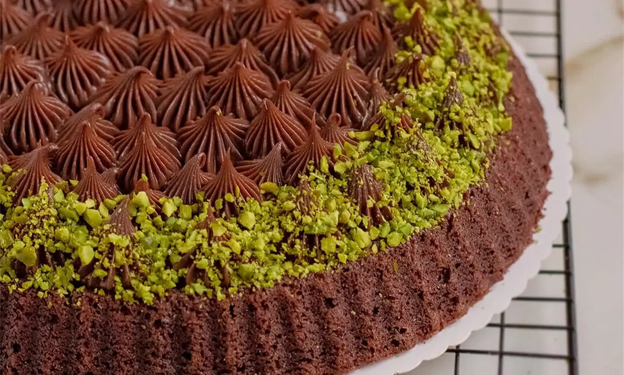 Kulatý čokoládový koláč s vroubkovaným okrajem, zdobený tmavými čokoládovými krémovými kopečky a vrstvou jemně nasekaných zelených pistácií