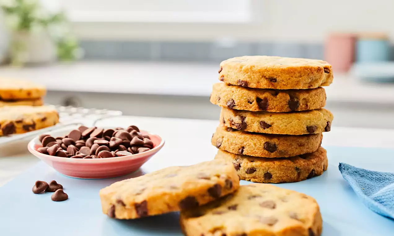 Golden round chocolate chip shortbread cookies stacked and scattered on a blue surface with a pink bowl of chocolate chips nearby