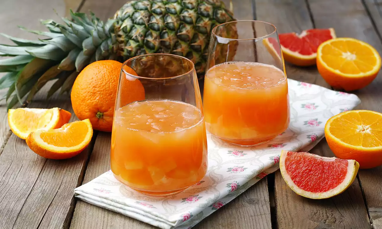 Cam bardaklarda taze sıkılmış greyfurt, portakal ve ananas parçaları içeren meyve suyu, pastel desenli bir örtü üzerinde sunulmuş