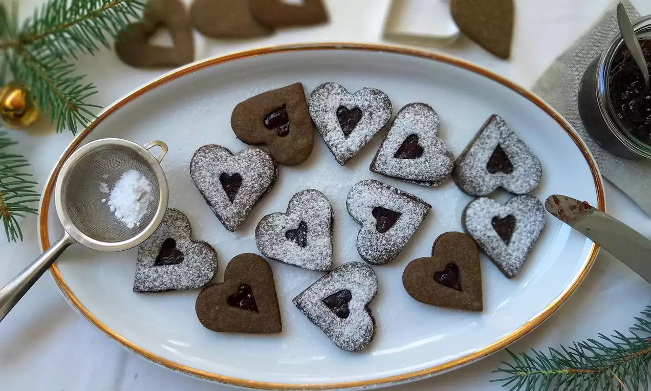 Maková srdíčka tmavohnědá a posypaná moučkovým cukrem, srdcovitý otvor plný rudé marmelády na podlouhlém bílém talíři vedle větvičky jedle