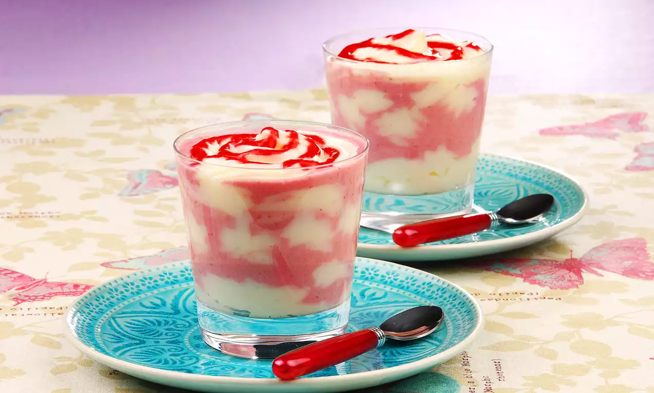 İnce cam bardaklarda, kremsi beyaz ve çilekli pembe katmanlarla sunulan tatlı, üzeri çilek sosuyla süslenmiş, mavi desenli tabaklarda