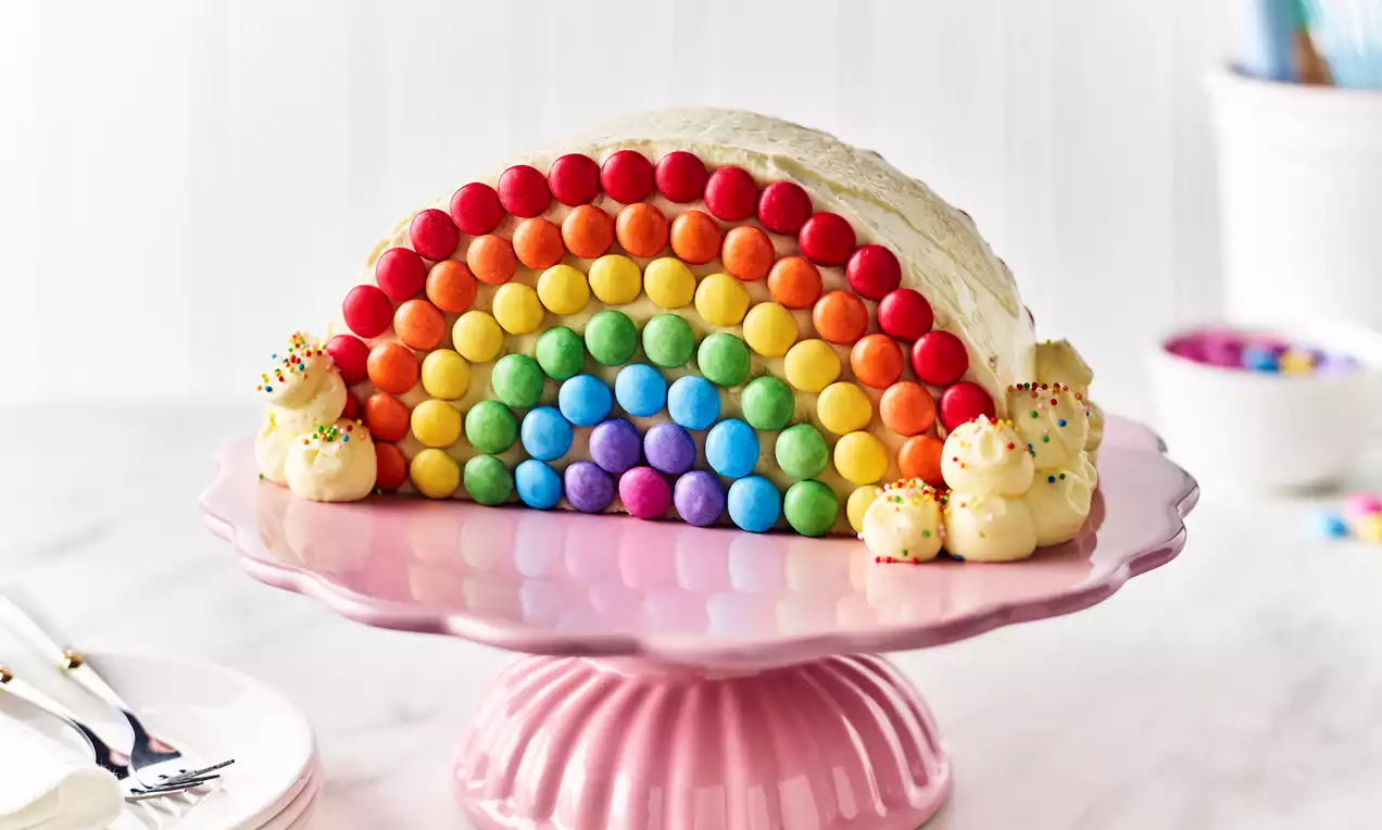 A semi-circular cake with rows of rainbow-coloured Smarties on white frosting, topped with cream swirls on a pink pedestal plate