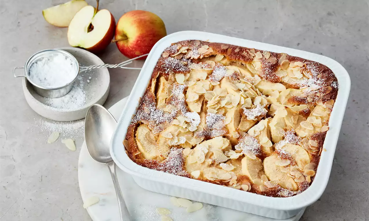 Goldbrauner Apfelauflauf mit zarten Mandelblättchen, leicht mit Puderzucker bestäubt, serviert in einer weißen Auflaufform auf hellem Untergrund