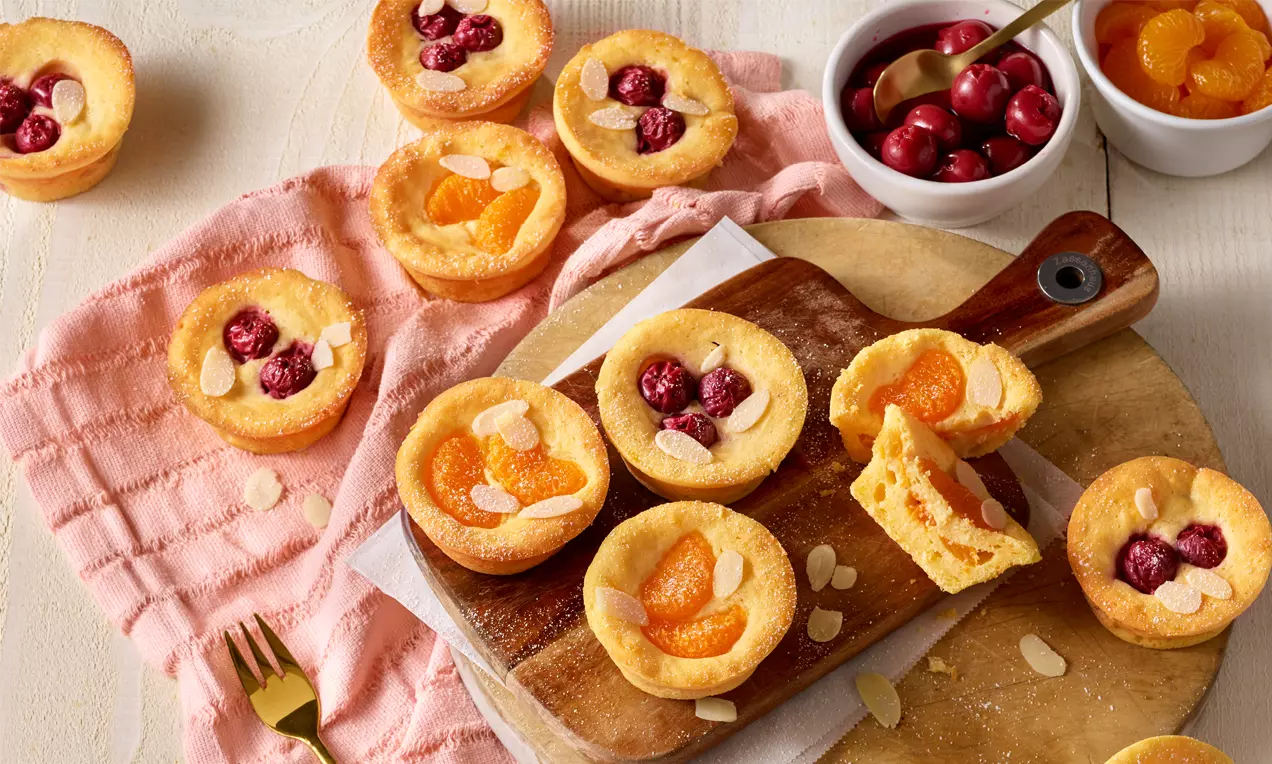 Kleine goldgelbe Topfenmuffins mit saftigen Mandarinen und Kirschen, dekoriert mit feinen Mandelblättchen auf einem Holzbrett angerichtet