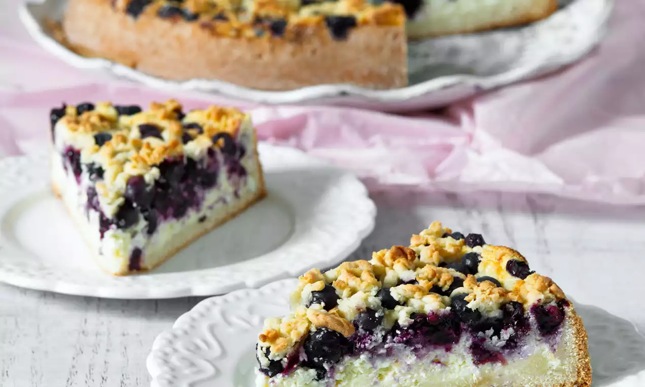 Kousek koláče s lehkou krémovou náplní, čerstvými borůvkami a drobenkou na zlatohnědém korpusu servírovaný na bílém zdobeném talíři