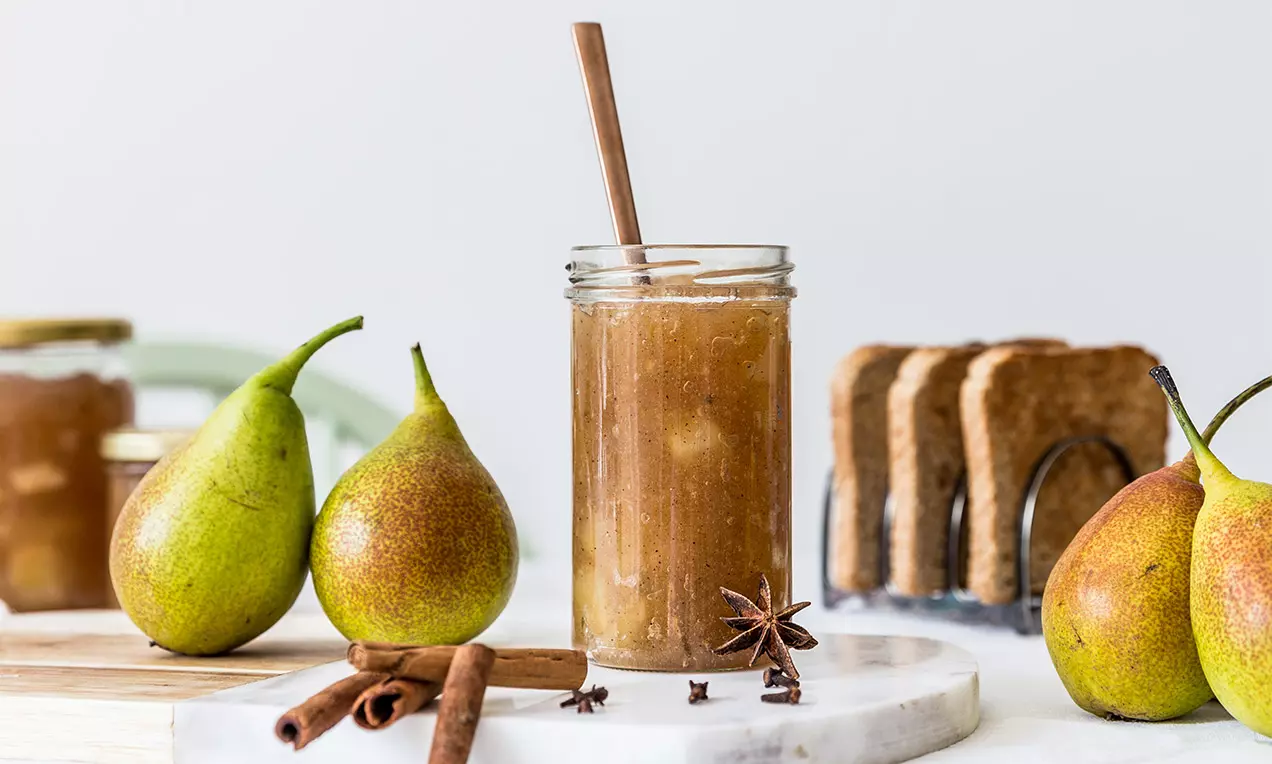 Jemne hnedý džem s viditeľnými kúskami hrušiek v sklenenom pohári s drevenou lyžičkou, obklopený hruškami, škoricou a toastmi na mramorovej doske