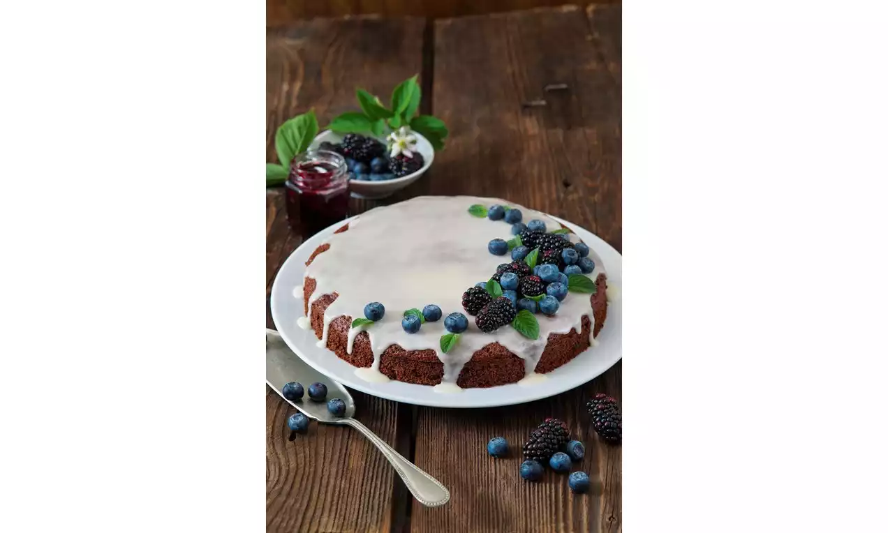 Perníkový dort kulatý, tmavě hnědý s bílou lesklou marcipánovou polevou, ozdobený ostružinami, borůvkami a lístky máty na dřevěném stole