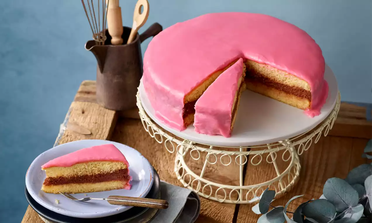 Punschtorte mit rosafarbenem Zuckerguss, hellen Biskuitschichten und rotem Füllungskern auf weißem Tortengitter mit Holz-Dekor