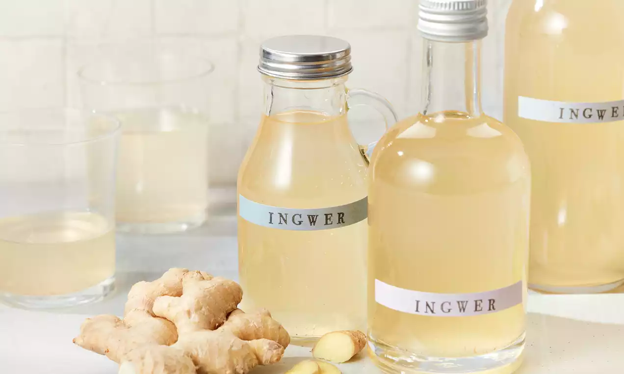 Hellgelber Ingwersirup in klaren Glasflaschen mit Etikett, daneben frische Ingwerknollen und Scheiben auf hellem Untergrund