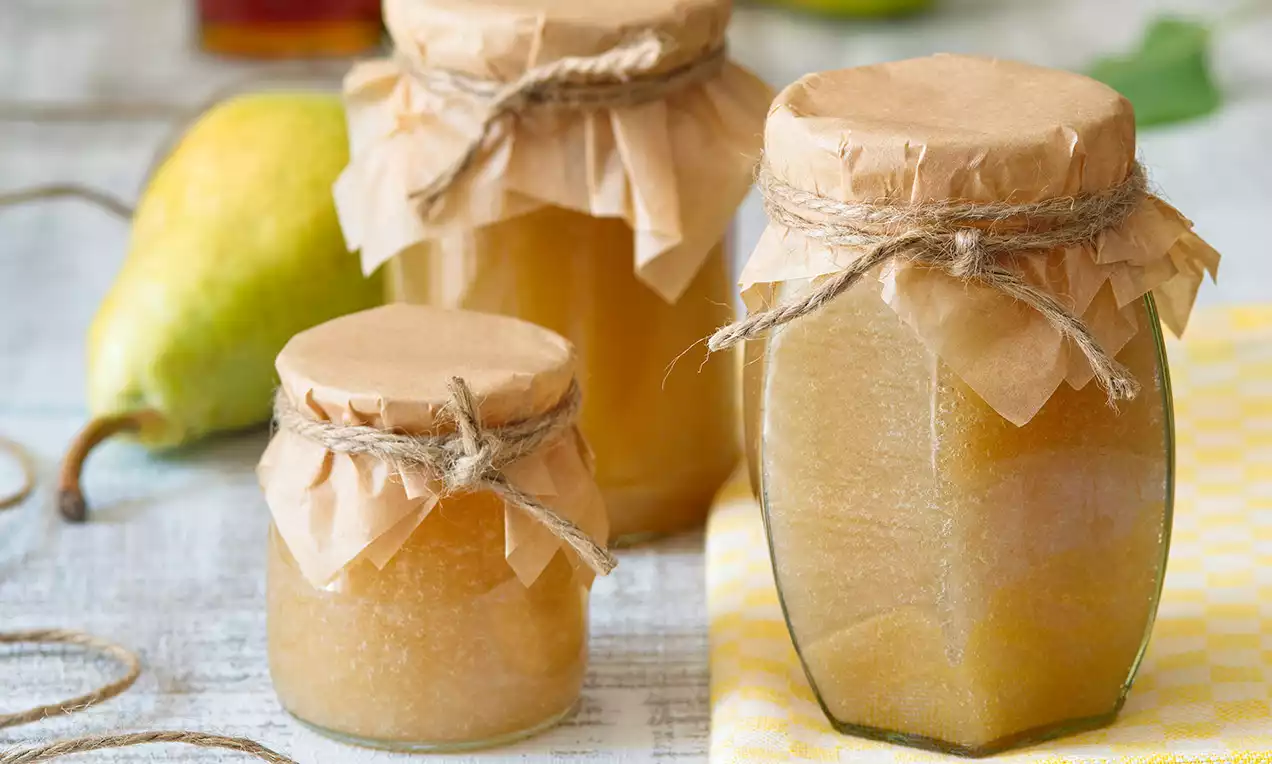 Sklenice hruškového džemu ve zlatavých tónech, zavázané přírodním provázkem, stojící na světlém povrchu vedle čerstvé hrušky
