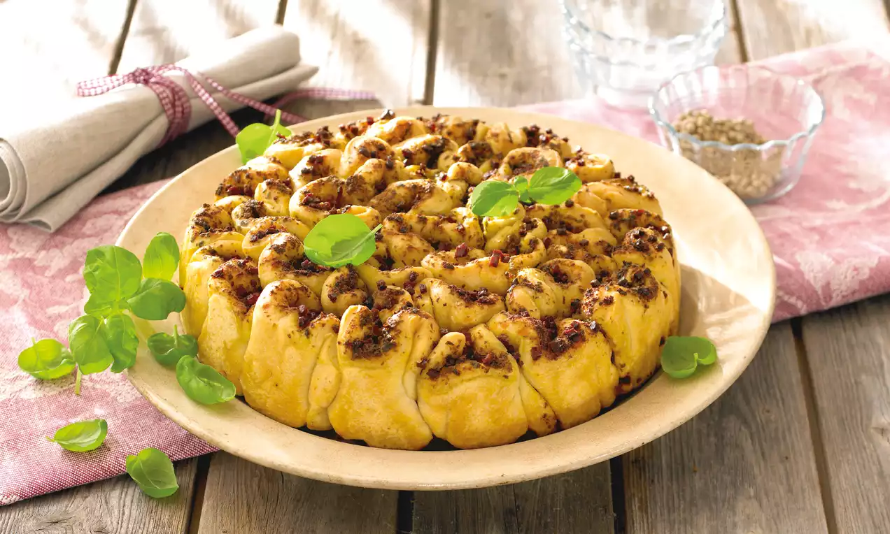 Goldbraunes Pestobrot in einer runden Form, spiralartig gefüllt mit Schinken und Pesto, garniert mit frischen Basilikumblättern auf einem Teller