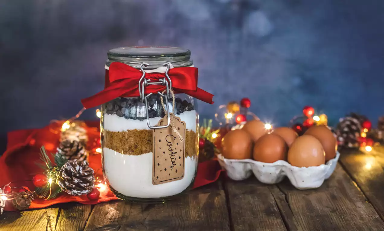 Mezcla de cookies en tarro de cristal decorado con lazo rojo y etiqueta, capas de ingredientes visibles junto a huevos y adornos navideños