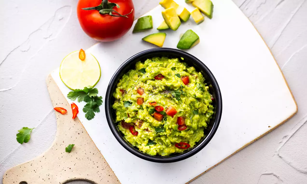 Guacamole vert vibrant dans un bol noir, garni de morceaux de tomate rouge et piment, entouré d’avocat, tomate et coriandre