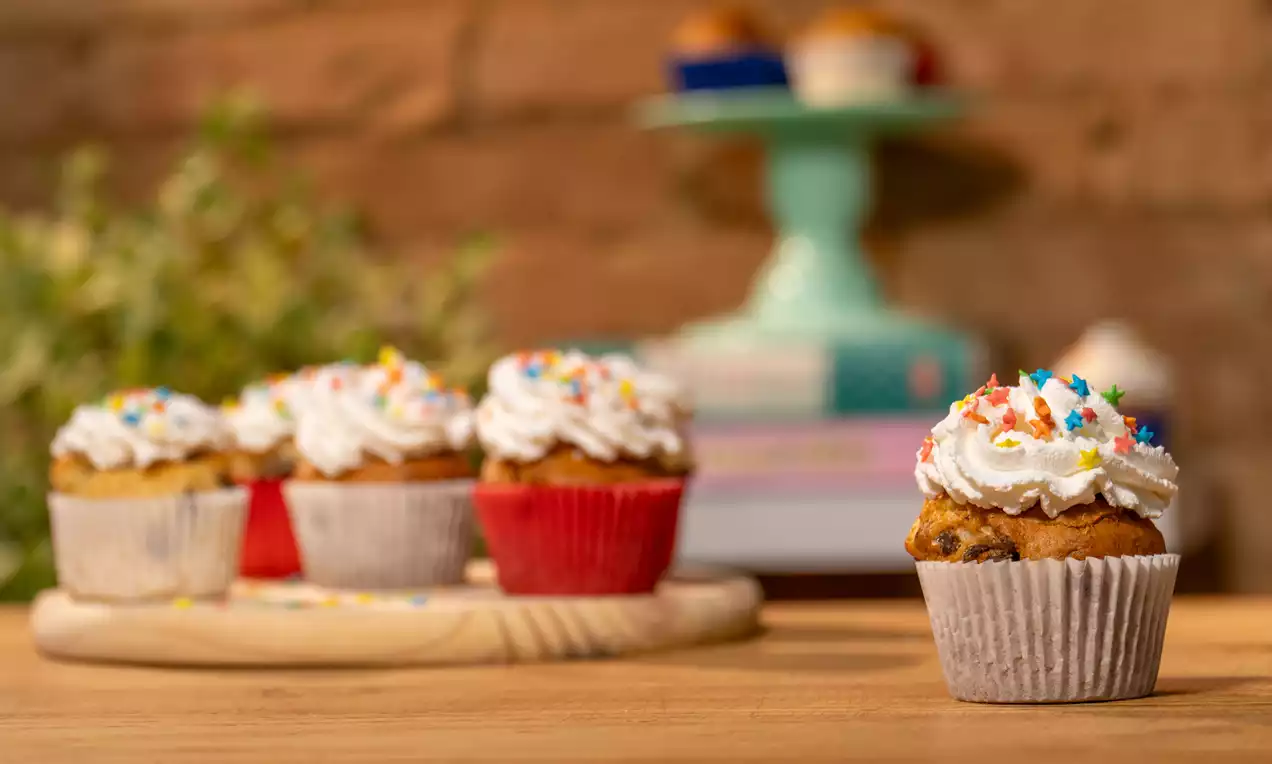 Muffin dourado com gotas de chocolate, cobertura de chantilly branco macio e confeitos coloridos, em forminha de papel listrada sobre mesa de madeira
