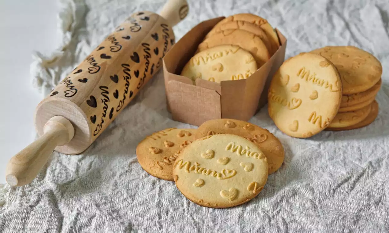 Světle zlatavé kulaté sušenky s jemnými otisky vzorů srdíček a nápisů uspořádané vedle hnědé krabičky a dekorativního válečku na lněném ubrusu