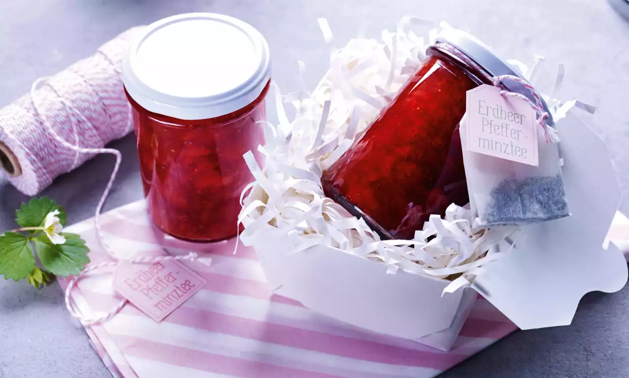 Ein Glas tiefrote Erdbeer-Pfefferminztee-Konfitüre liegt schräg in weißem Papiernest, daneben rosa Etikett und Stoffbeutelchen.