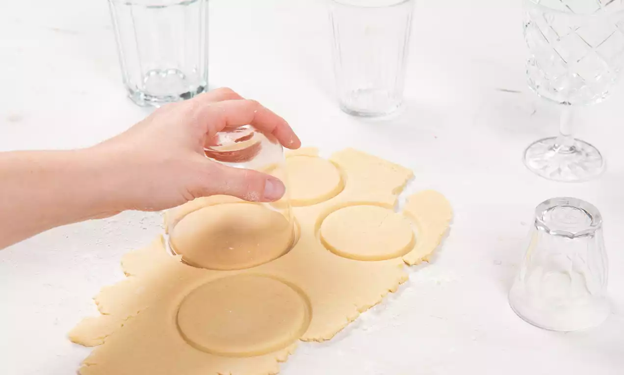 Hand, die mit einem Glas Kreise aus einem hellen, glatten Teig aussticht, auf einer weißen Arbeitsfläche mit verstreuten Gläsern