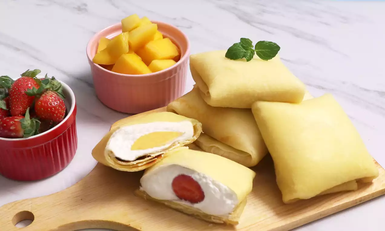 Golden crepes folded into rectangles on a wooden board with visible white cream fillings and fruit, served with strawberry and mango sides