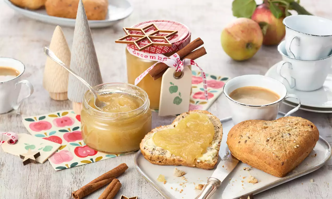 Apfel-Zimt-Marmelade in einem offenen Glas mit goldgelber Farbe neben einer Scheibe Brot, Zimtstangen und Dekoration auf hellem Holz