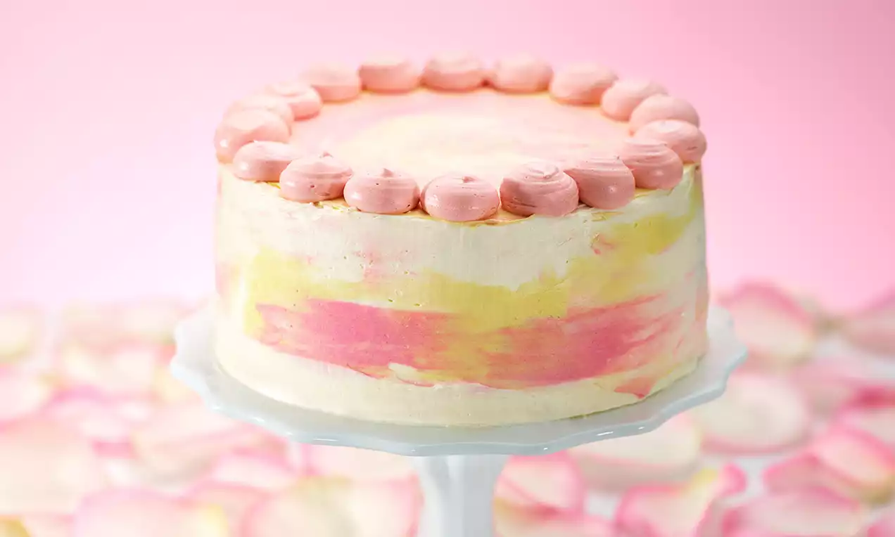 Torta al cioccolato con superficie sfumata giallo e rosa, decorata con ciuffi di crema rosa su un’alzata bianca e petali sullo sfondo