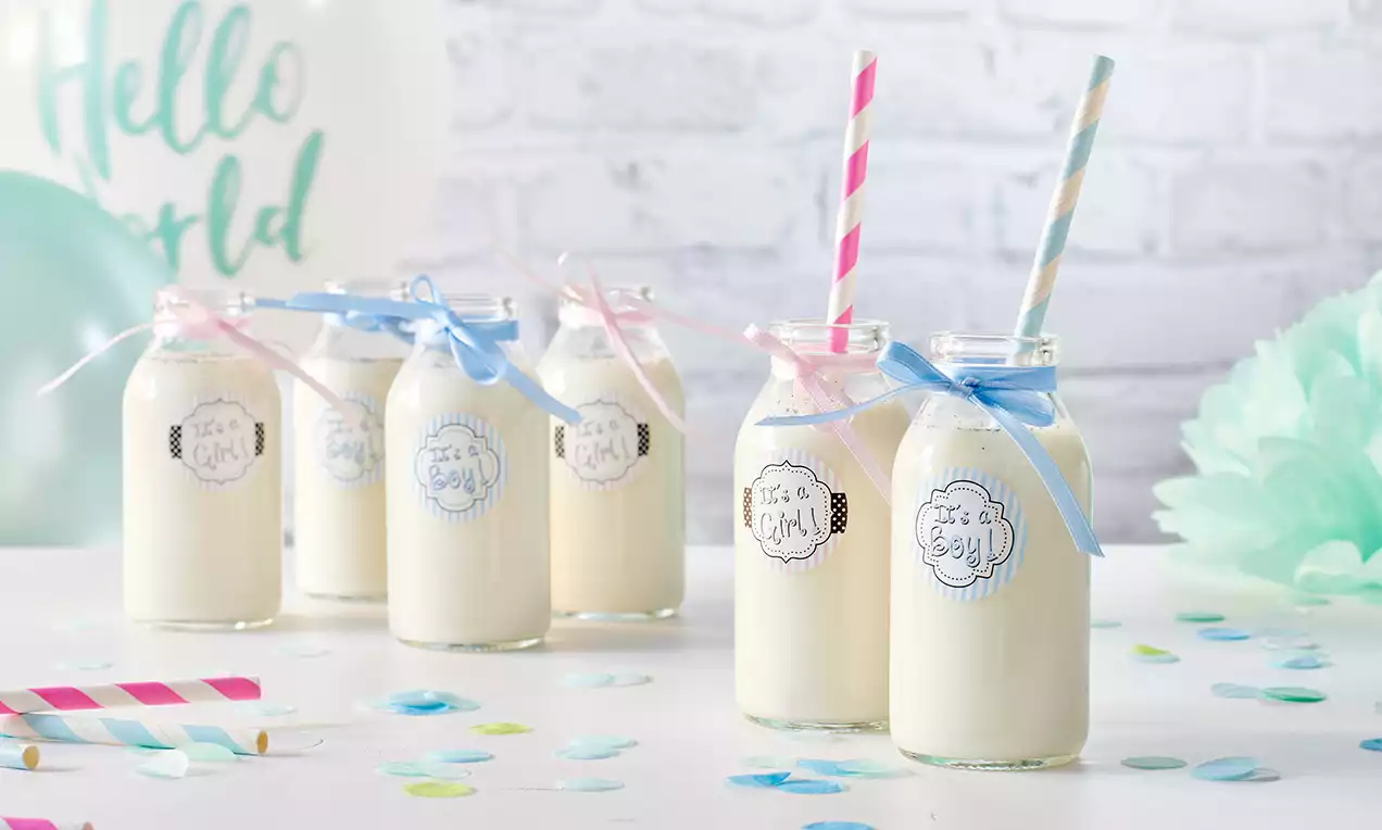 Vanillemilch in kleinen Glasflaschen mit pastellfarbenen Strohhalmen und Schleifen, auf einem weißen Tisch mit Konfetti und Dekoration arrangiert
