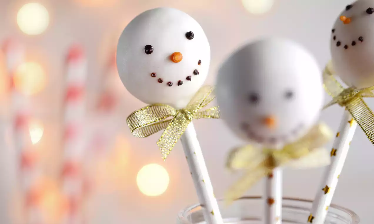 Białe lollipopsy ozdobione na kształt uśmiechniętych bałwanków z pomarańczowym noskiem, umieszczone na patyczkach z złotymi kokardkami