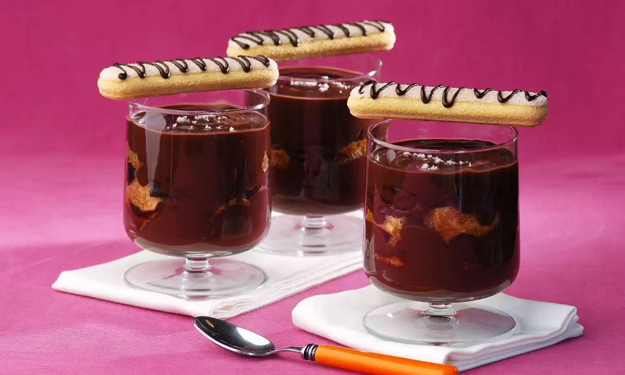 Cam bardaklarda yoğun çikolatalı krem, içinde bisküvi parçalarıyla katmanlı, üzerinde çikolata çizgili kedi dili bisküvi ile sunulmuş
