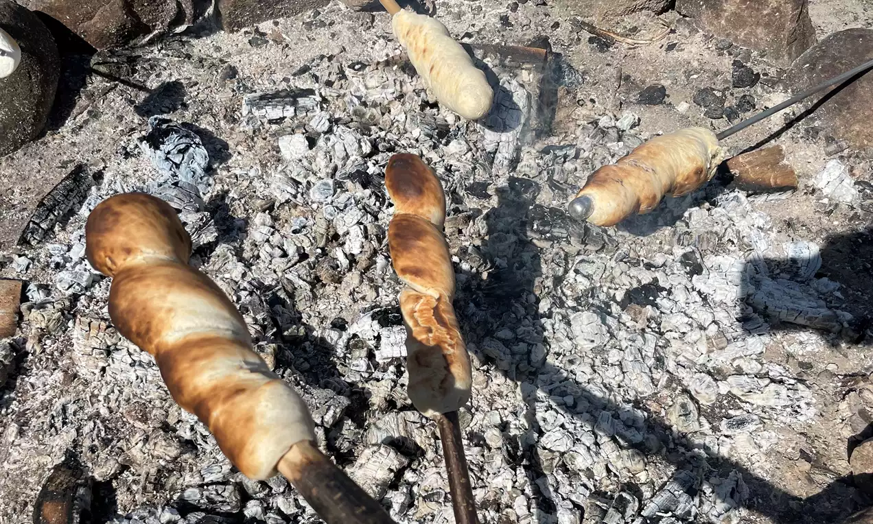 Snobrød med gyldne og lysebrune spiralformede skorper på pinde, ristet over glødende aske mellem spredte kul og sten
