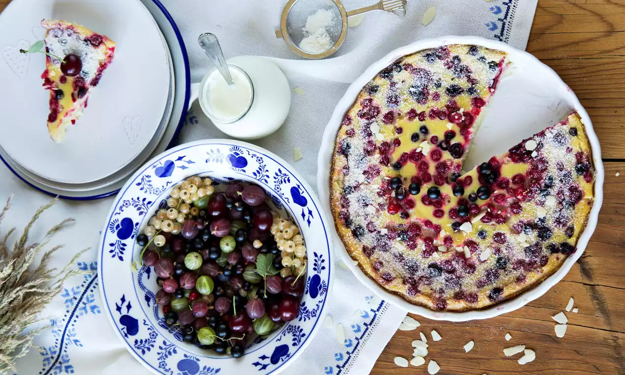 Clafoutis so zlatohnedými okrajmi a krémovým žltým stredom posiaty červenými a fialovými bobuľami, jemne pocukrovaný, na bielom obruse s dekoráciami