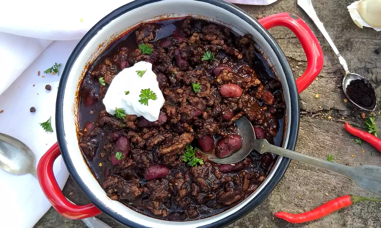 Hlboký čierny kovový hrniec s červenými rúčkami, naplnený tmavohnedým mäsovým ragú, červenými fazuľami, lyžicou smotany a čerstvou petržlenovou vňaťou