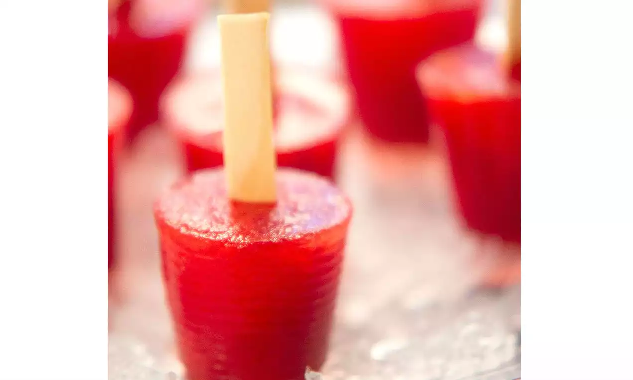 Picolés pequenos de tom vermelho vibrante com textura lisa e palitos de madeira clara dispostos sobre superfície translúcida com brilho