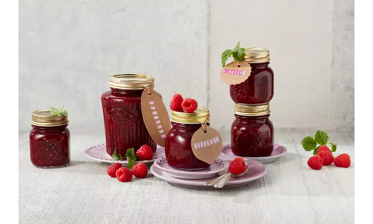 Himbeermarmelade in klaren Gläsern mit goldenen Deckeln, dekoriert mit Himbeeren, Minze und beschrifteten Anhängern auf lila Tellern