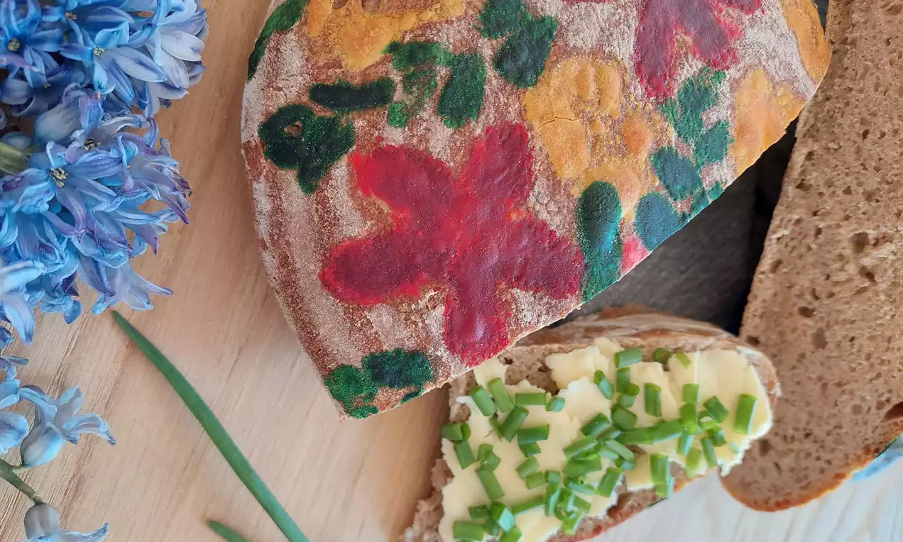 Žitný kváskový chlieb zdobený farebnými jarnými kvetmi, s krajcom potretým maslom a posypaným pažítkou na drevenej doske vedľa modrých kvetov