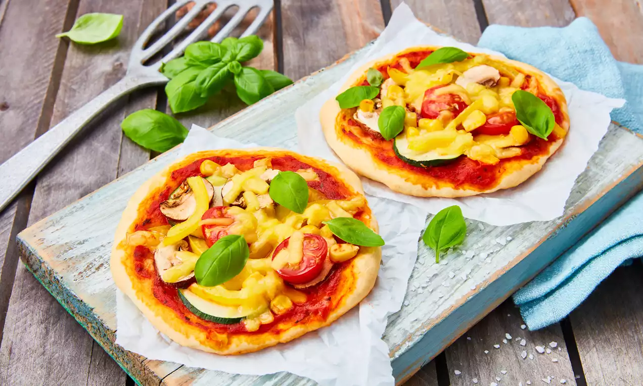 Zwei kleine, runde Pizzen mit goldbraunem Boden, roter Tomatensauce, frischem Gemüse, geschmolzenem Käse und grünem Basilikum auf Holzbrett