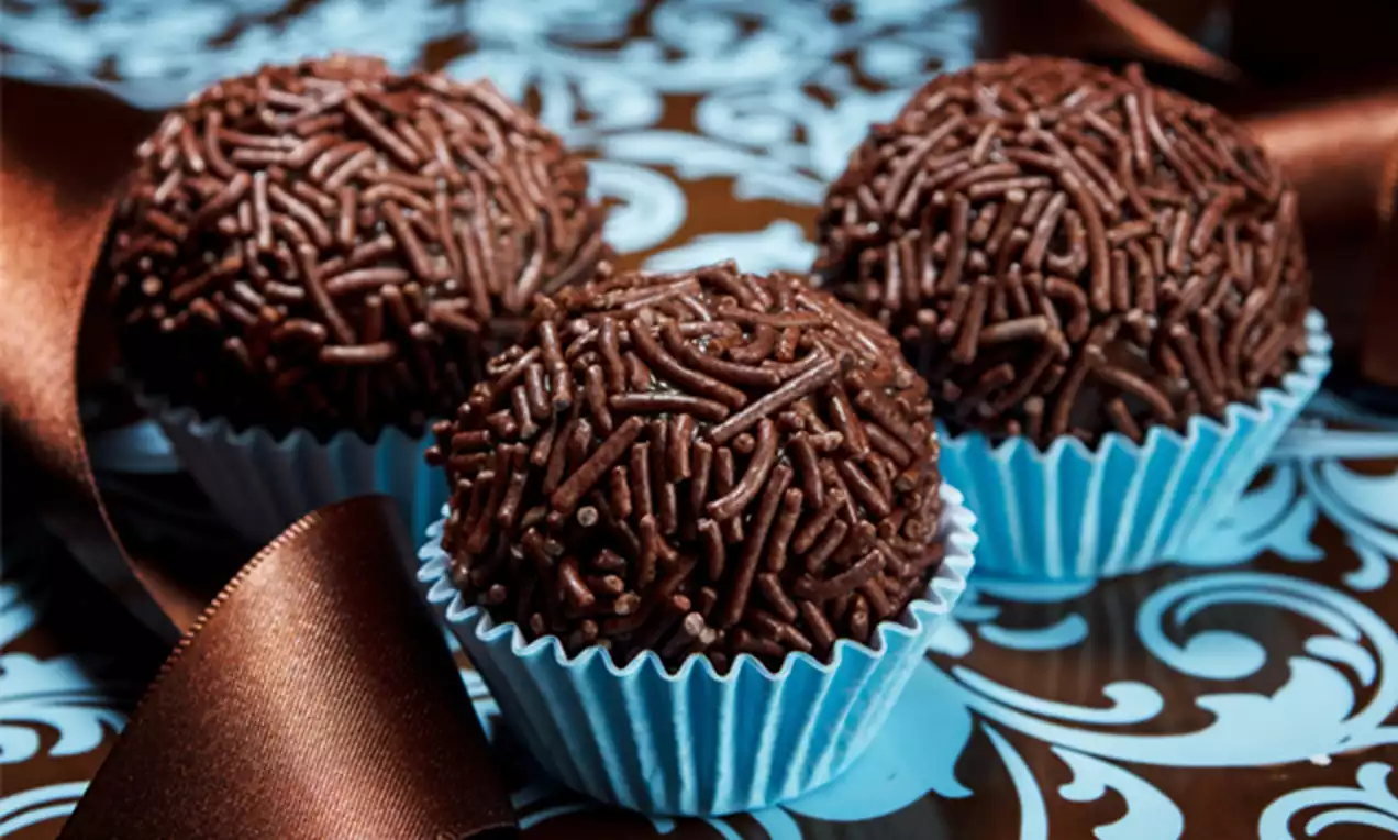 Three round chocolate truffles coated with dark brown sprinkles in blue paper cups on a patterned surface with a satin ribbon