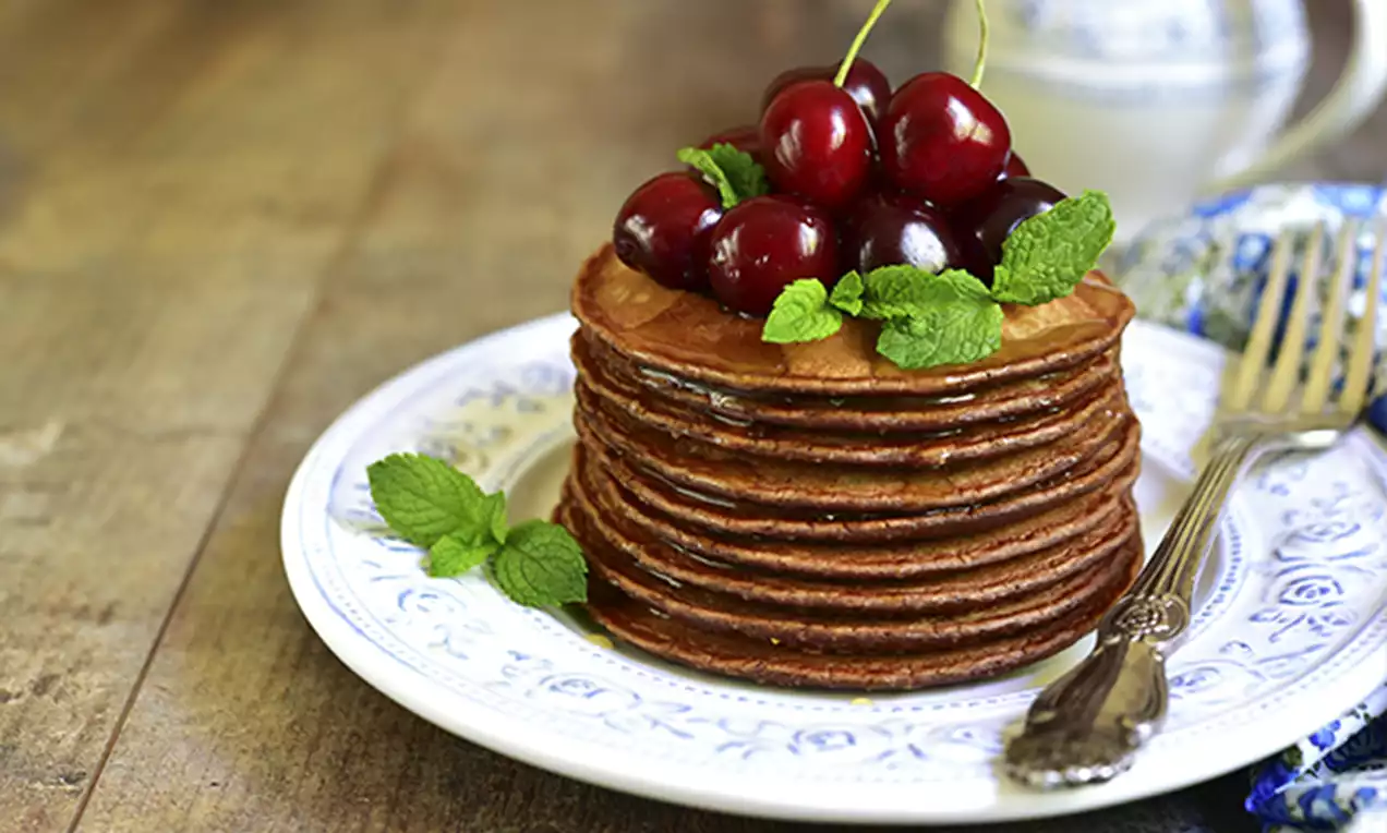 A stack of round chocolate pancakes with a glossy surface, topped with red cherries and fresh green mint leaves on a patterned white plate