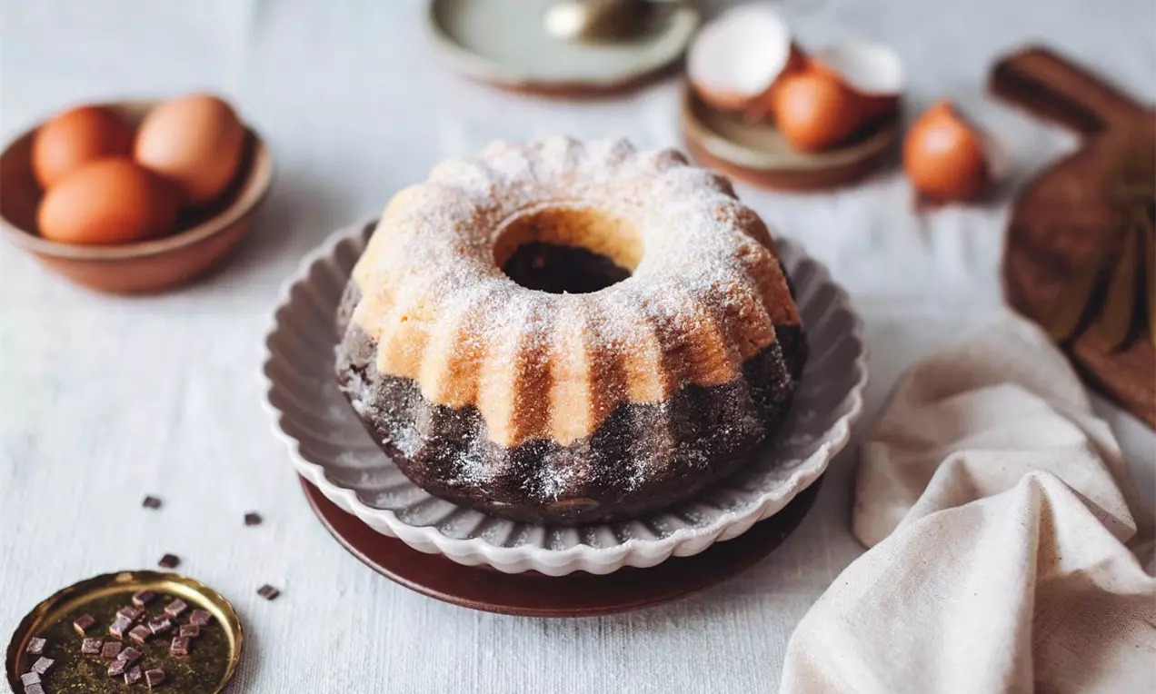 Bábovka zo svetlého a tmavého cesta posypaná práškovým cukrom na pracovnej ploche s vajíčkami a valčekom v pozadí.
