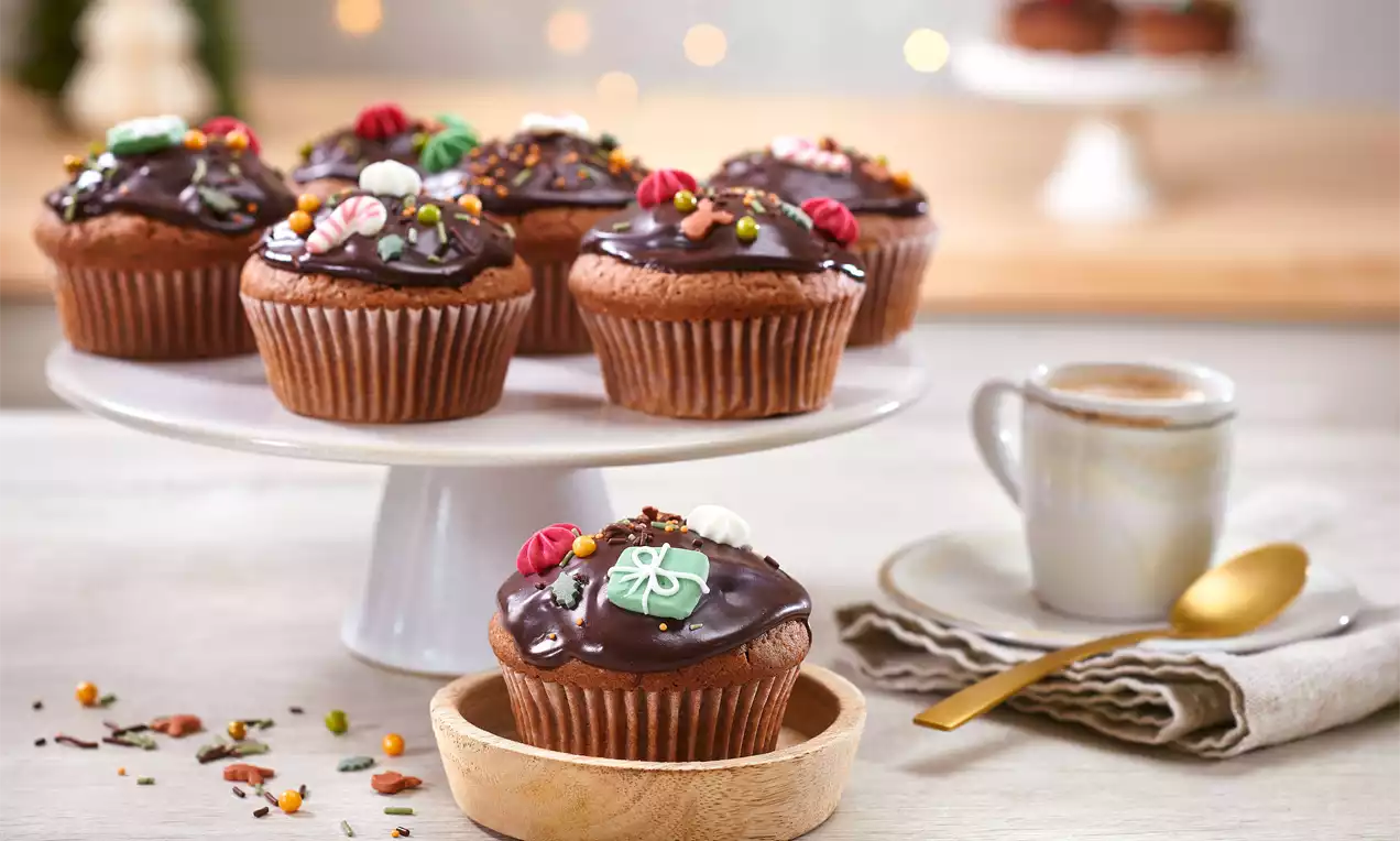 Gewürzmuffins mit glänzender Schokoladenglasur, dekoriert mit bunten Zuckerstreuseln und Figuren, arrangiert auf einem weißen Tortenständer