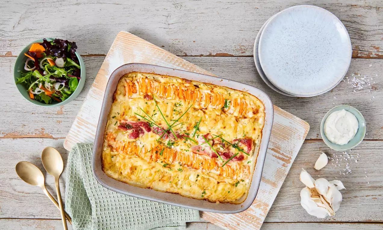 Kartoffel-Möhren-Auflauf in einer goldbraun gratinierten Oberfläche mit orangefarbenen Möhrenstreifen und Kräutern in einer weißen Auflaufform