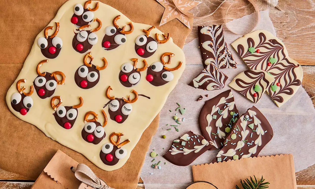 Weiße Schokolade mit Rentier-Verzierung aus roten Nasen, Augen und Salzbrezeln neben marmorierten und bestreuten braunen Schokoladenstücken