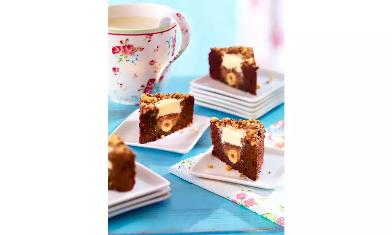 Tamnosmeđi kutni brownies s kremastim bijelim punjenjem i lješnjacima, posluženi na bijelim tanjurićima uz šarenu cvjetnu šalicu na plavom stolu