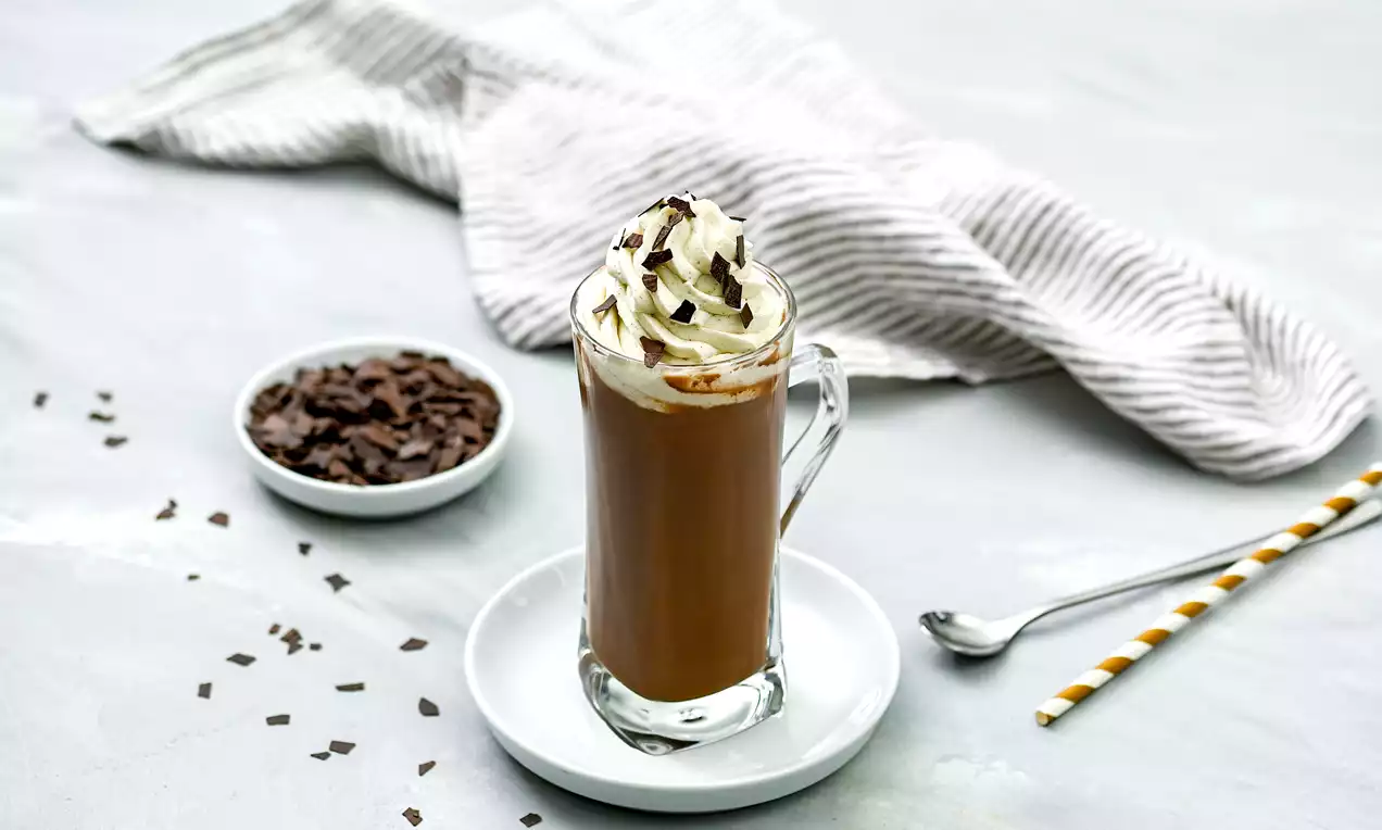 Glas mit braunem Kakao, getoppt mit weißer Sahne und dunklen Schokoraspeln, auf einer weißen Untertasse mit Streifenstrohhalm drapiert