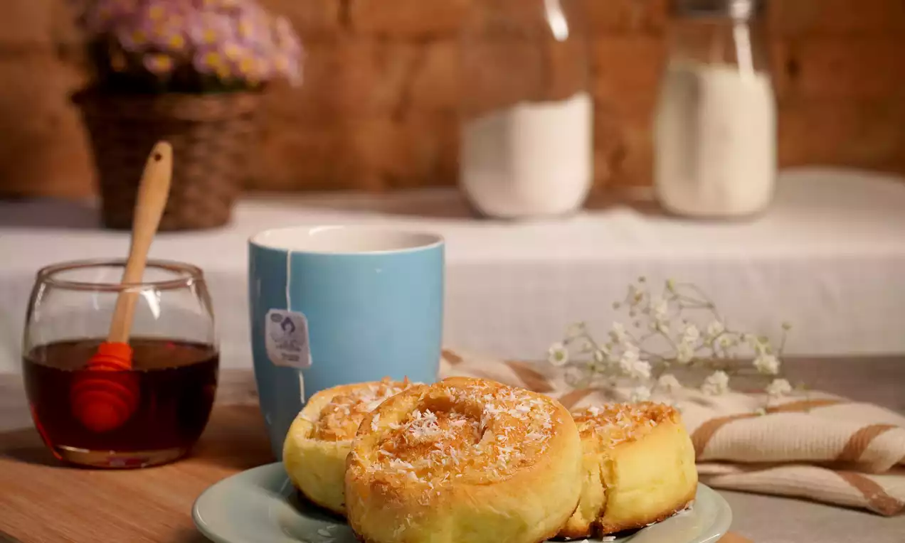 Três rolos dourados e macios de massa em espiral polvilhados com coco, dispostos em um prato azul claro ao lado de uma xícara azul e mel