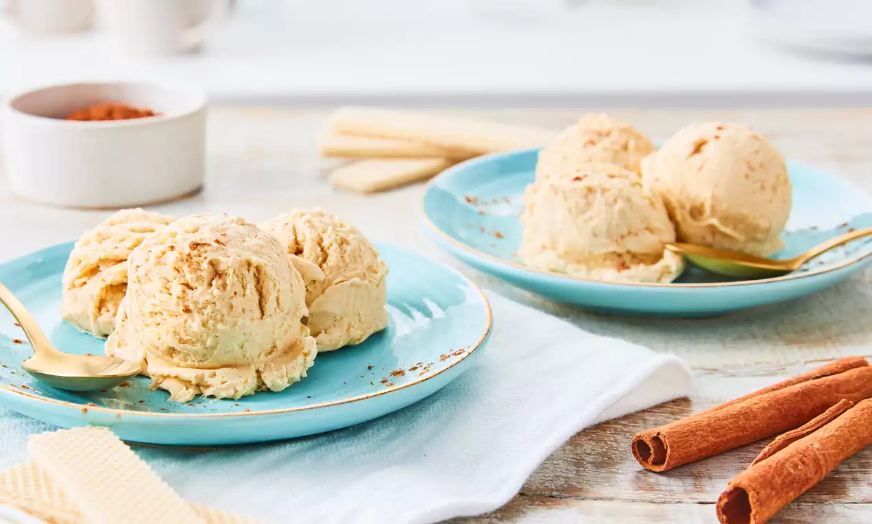 Zimteis in hellbeigen, cremigen Kugeln in einer Schale, mit braunem Zimtpulver bestreut, dazu eine dekorative Zimtstange und ein Holzlöffel